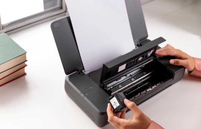 Person installing an ink cartridge into a portable printer on a desk beside a stack of books